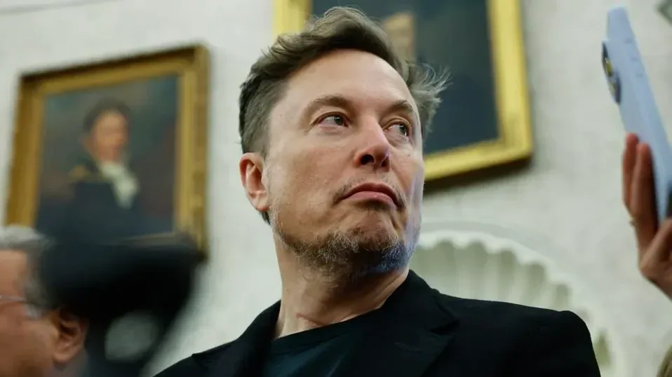Retrato de Elon Musk vistiendo traje oscuro con cuadros al fondo y una mano sosteniendo un teléfono móvil.