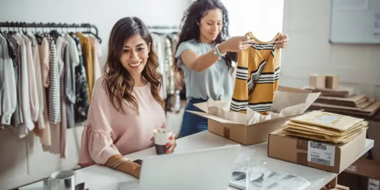 Dos mujeres gestionan un negocio de moda online. Una usa un portátil, la otra empaqueta ropa en cajas para envío. Fondo con percheros.