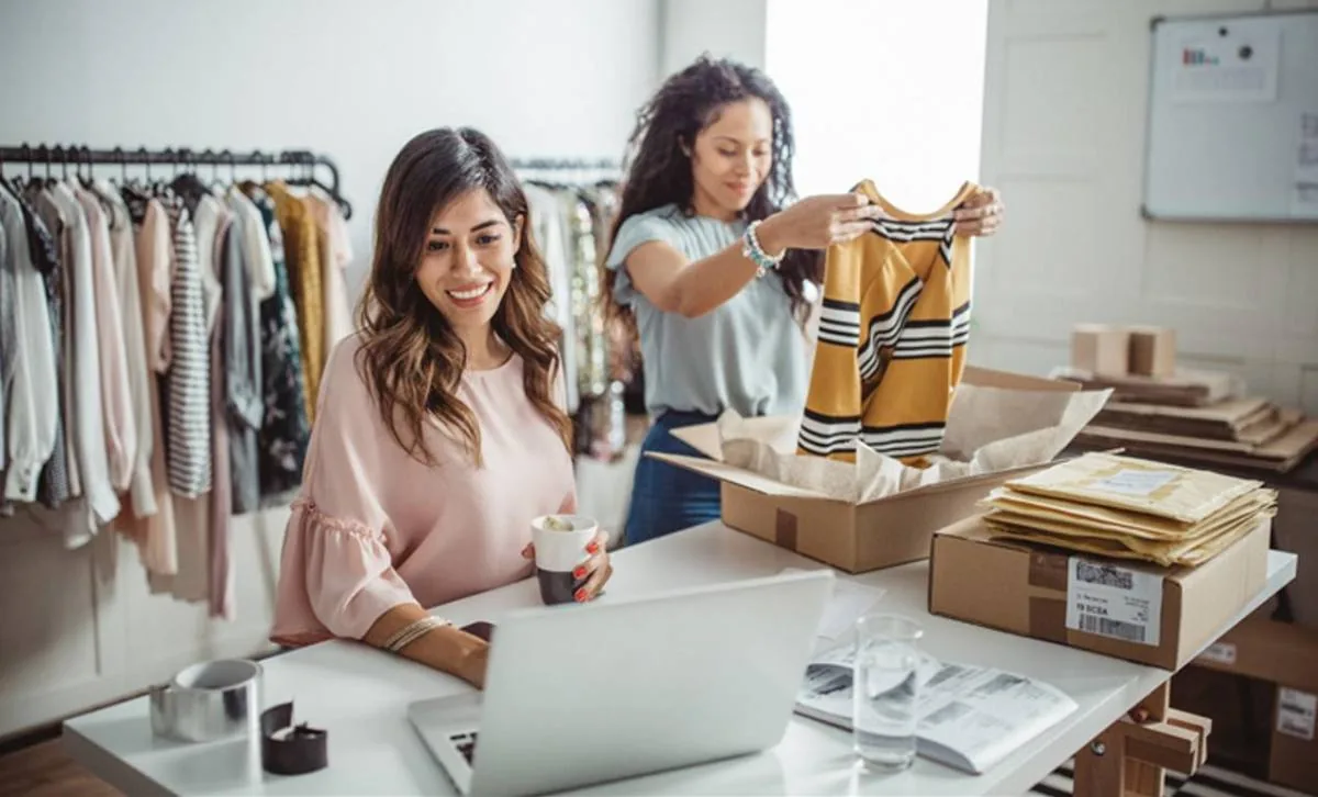 Dos mujeres gestionan un negocio de moda online. Una usa un portátil, la otra empaqueta ropa en cajas para envío. Fondo con percheros.