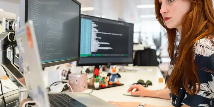 Mujer pelirroja trabajando frente a dos monitores de computadora con código de programación en un escritorio de oficina.