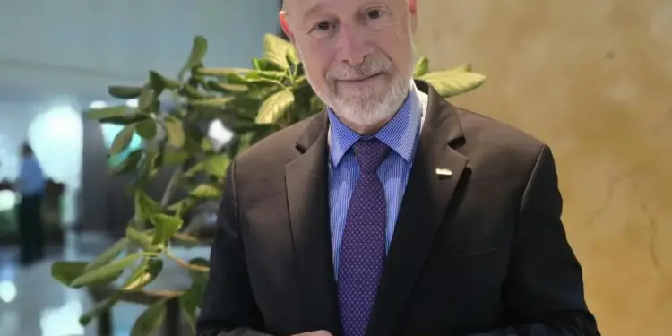 Retrato de un hombre caucásico de mediana edad, calvo, con barba gris, vestido con traje negro, camisa azul a rayas y corbata morada a lunares, sonriendo a la cámara.