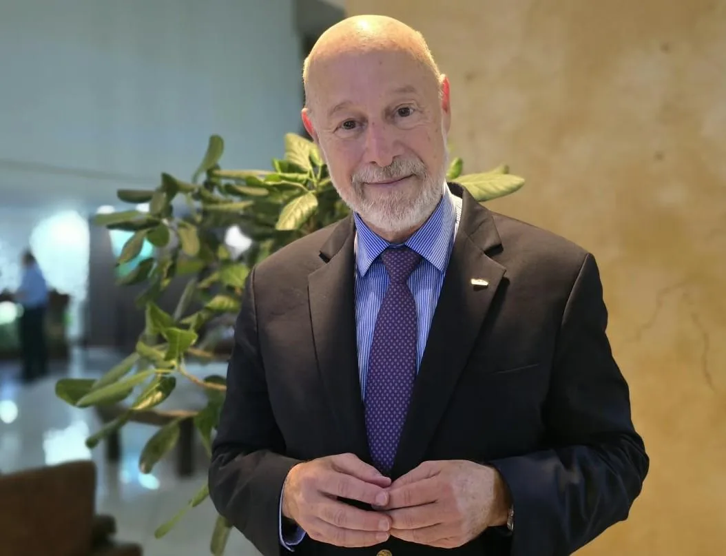 Retrato de un hombre caucásico de mediana edad, calvo, con barba gris, vestido con traje negro, camisa azul a rayas y corbata morada a lunares, sonriendo a la cámara.
