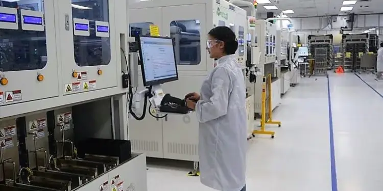 Una mujer con bata de laboratorio y gafas de seguridad opera una computadora en una línea de producción industrial, rodeada de maquinaria.