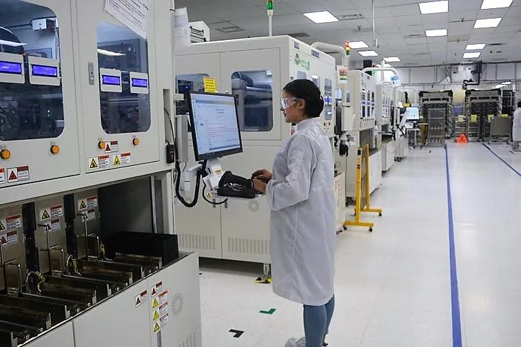 Una mujer con bata de laboratorio y gafas de seguridad opera una computadora en una línea de producción industrial, rodeada de maquinaria.