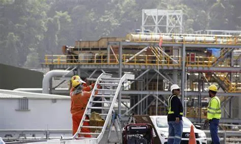 Trabajadores con equipo de seguridad en una planta industrial con estructuras metálicas, tuberías y maquinaria. Un operario sube escaleras, otros dos conversan junto a un vehículo.