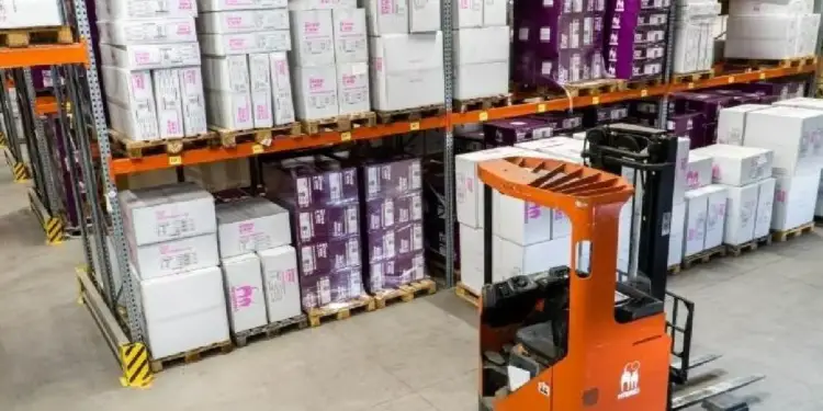 Vista de un almacén con estanterías metálicas llenas de cajas apiladas en palets, y una carretilla elevadora naranja en primer plano.
