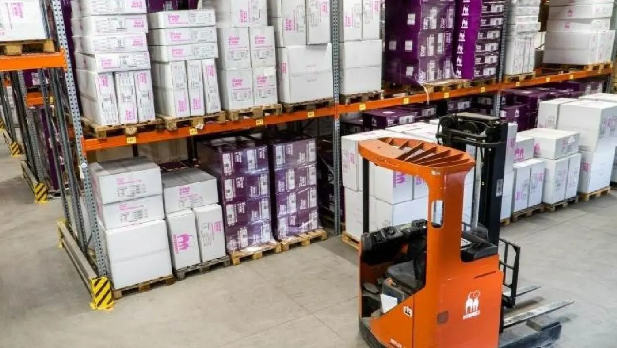 Vista de un almacén con estanterías metálicas llenas de cajas apiladas en palets, y una carretilla elevadora naranja en primer plano.