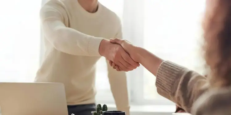 Dos profesionales estrechando manos en una oficina luminosa, simbolizando un acuerdo o colaboración. Hay un portátil y tazas de café en la mesa.
