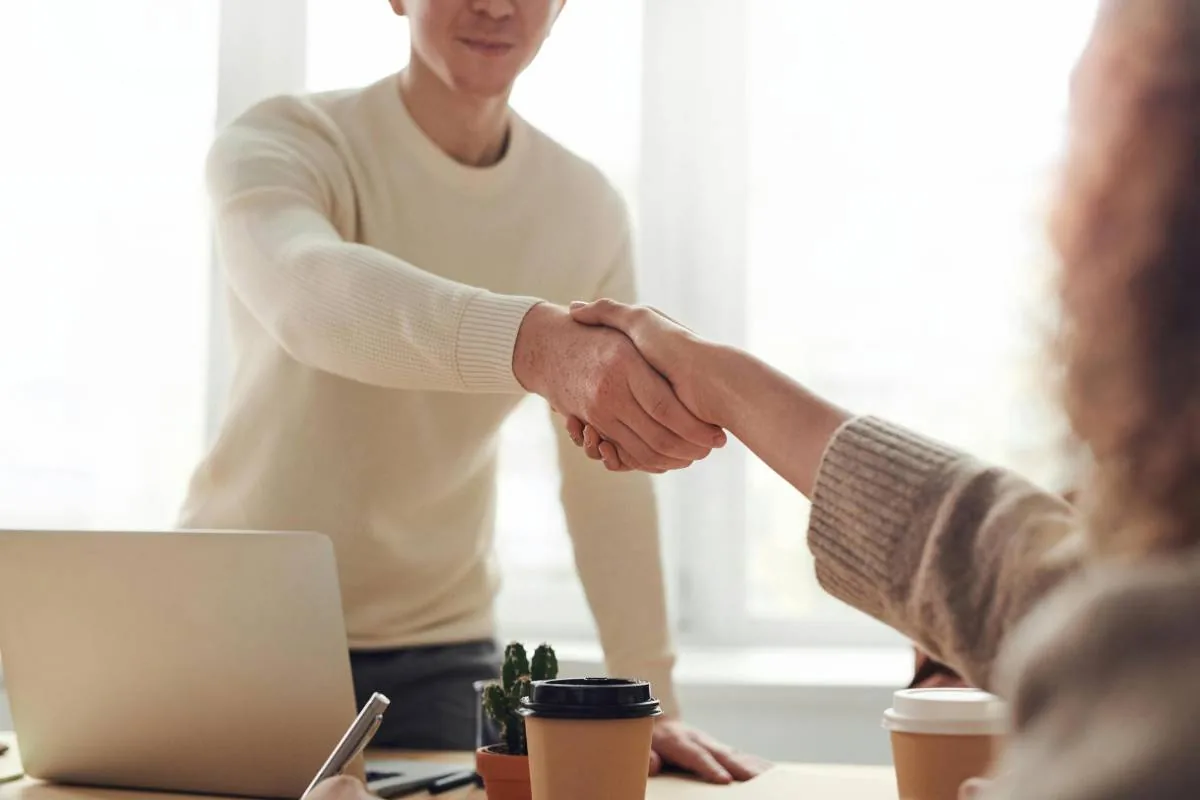 Dos profesionales estrechando manos en una oficina luminosa, simbolizando un acuerdo o colaboración. Hay un portátil y tazas de café en la mesa.