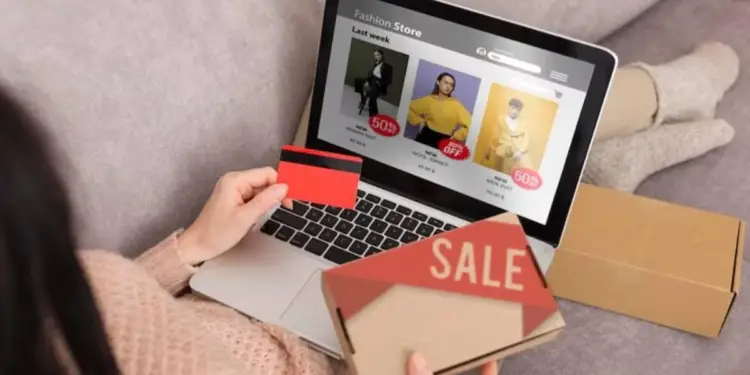 Persona comprando online con tarjeta de crédito en laptop, viendo ofertas de moda. Sostiene una caja de "SALE" y otra de envío.