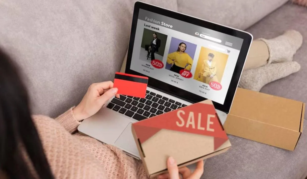 Persona comprando online con tarjeta de crédito en laptop, viendo ofertas de moda. Sostiene una caja de "SALE" y otra de envío.