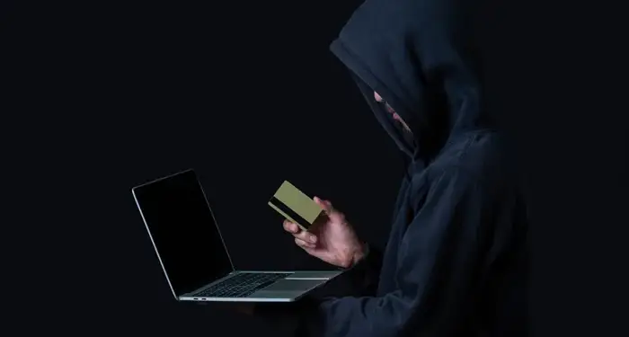 Persona con capucha negra usando una laptop y sosteniendo una tarjeta de crédito sobre fondo oscuro.