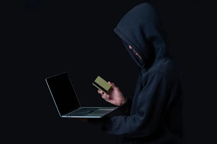 Persona con capucha negra usando una laptop y sosteniendo una tarjeta de crédito sobre fondo oscuro.