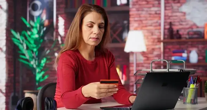 Mujer de mediana edad con blusa roja sentada frente a un portátil negro, sosteniendo una tarjeta de crédito naranja y negra. Realiza una compra o transacción online en un entorno de oficina o estudio.