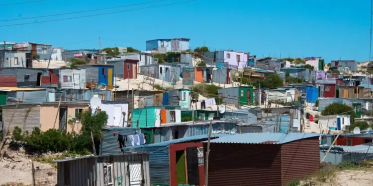 Vista panorámica de un asentamiento informal con numerosas viviendas precarias de chapa metálica en un terreno arenoso bajo un cielo azul claro. Refleja desafíos socioeconómicos.