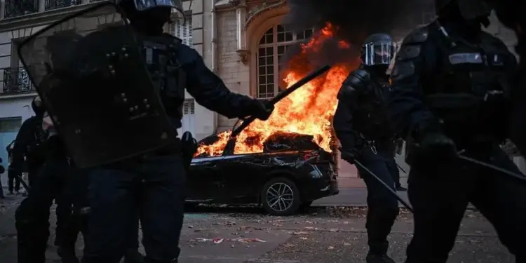 Agentes policiales antidisturbios patrullando frente a un automóvil en llamas durante disturbios urbanos.