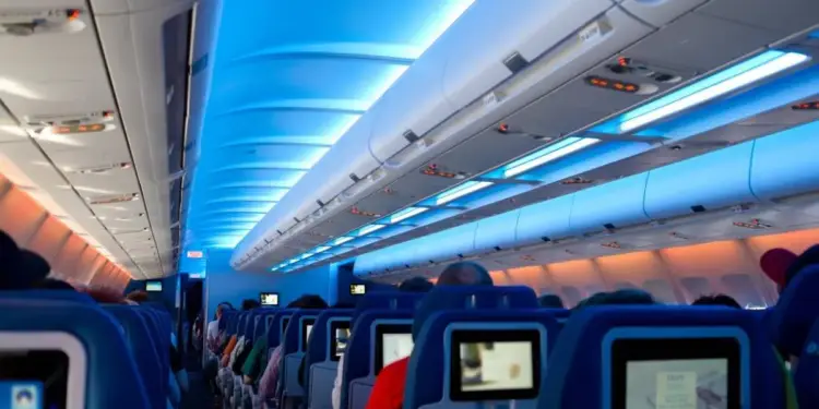 Interior de un avión con asientos azules y pantallas de entretenimiento, iluminado con luces LED azules y naranjas en el techo.