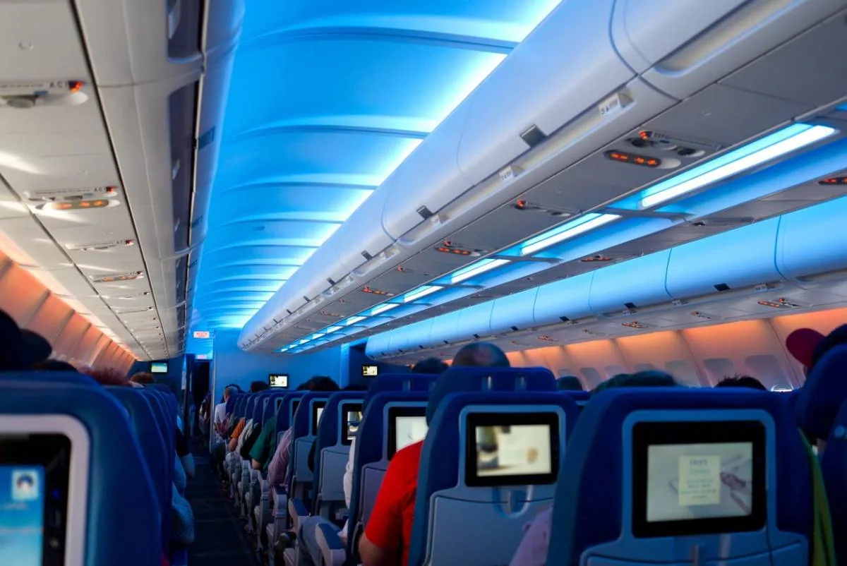 Interior de un avión con asientos azules y pantallas de entretenimiento, iluminado con luces LED azules y naranjas en el techo.