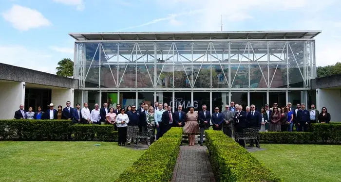 Amplio grupo de hombres y mujeres de negocios posando frente a un moderno edificio de cristal y arquitectura contemporánea.