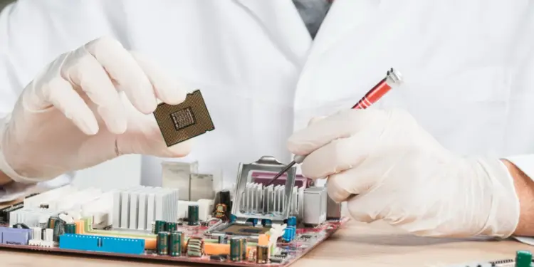 Técnico con bata blanca y guantes manipula un microprocesador y una placa base con herramientas.