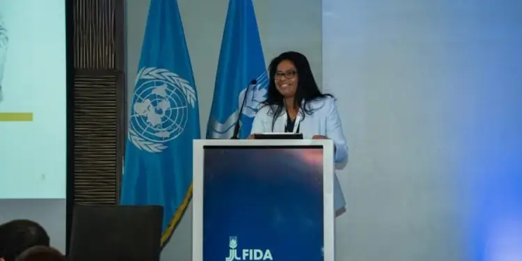 Mujer de pelo oscuro sonriendo mientras habla desde un atril con el logotipo de FIDA y banderas azules al fondo
