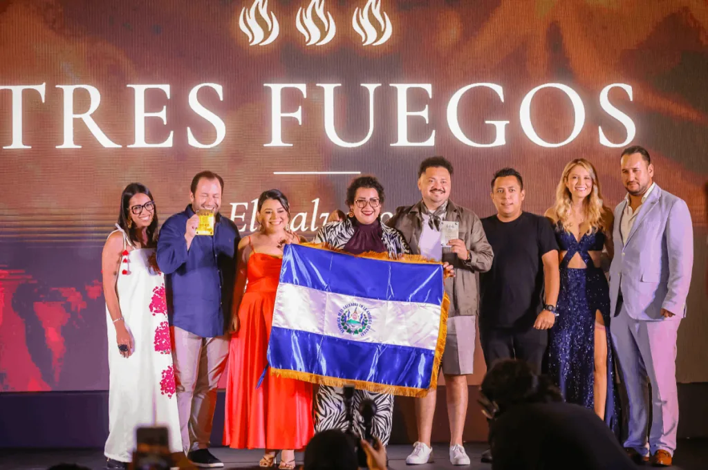 Grupo de ocho personas posando en un escenario frente a un cartel que dice TRES FUEGOS, sosteniendo una bandera y premios.
