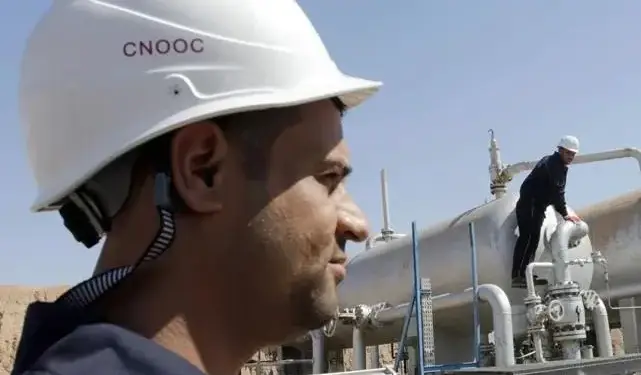 Hombre con casco de seguridad CNOOC y uniforme azul en entorno industrial. Otro trabajador supervisa tuberías y tanques.