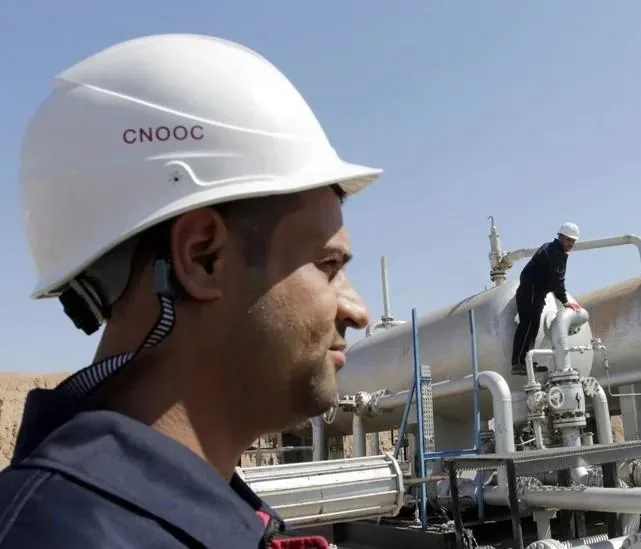 Hombre con casco de seguridad CNOOC y uniforme azul en entorno industrial. Otro trabajador supervisa tuberías y tanques.