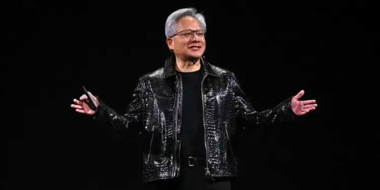 Hombre con cabello gris y gafas, chaqueta negra brillante texturizada, presentando en escenario oscuro. Sostiene un control remoto y gesticula con las manos abiertas.