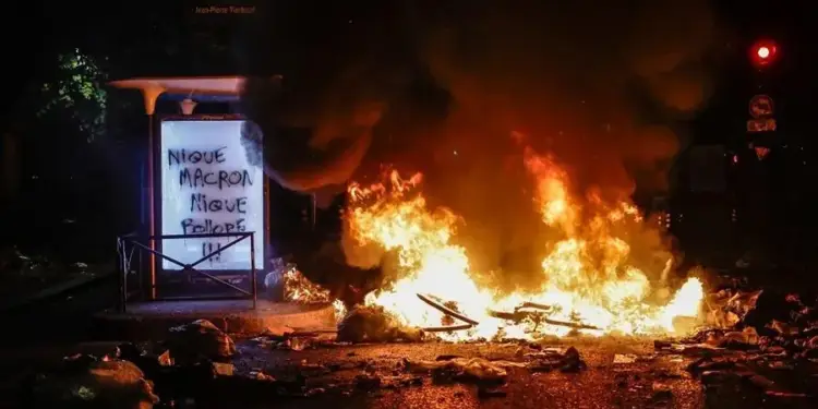 Gran hoguera con fuego intenso y humo negro en la calle durante la noche, junto a una parada de autobús iluminada.