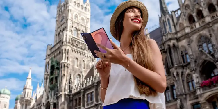Una mujer con sombrero de paja sostiene un teléfono móvil plegable frente a una arquitectura histórica de estilo gótico.