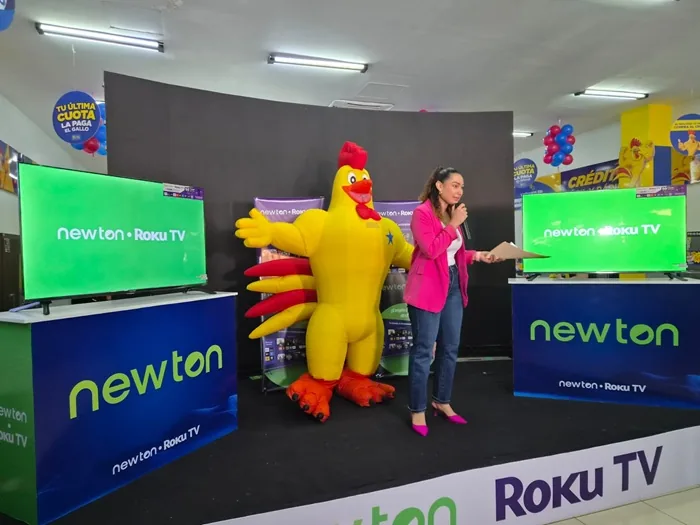 Mujer hablando con micrófono en un escenario junto a una botarga de gallo y dos pantallas de televisión.