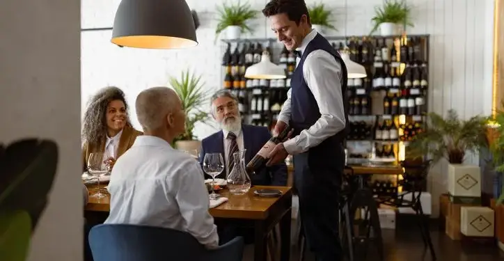 Camarero profesional presenta una botella de vino a comensales en una cena de negocios. Grupo de personas en un restaurante elegante con estanterías de vino al fondo.