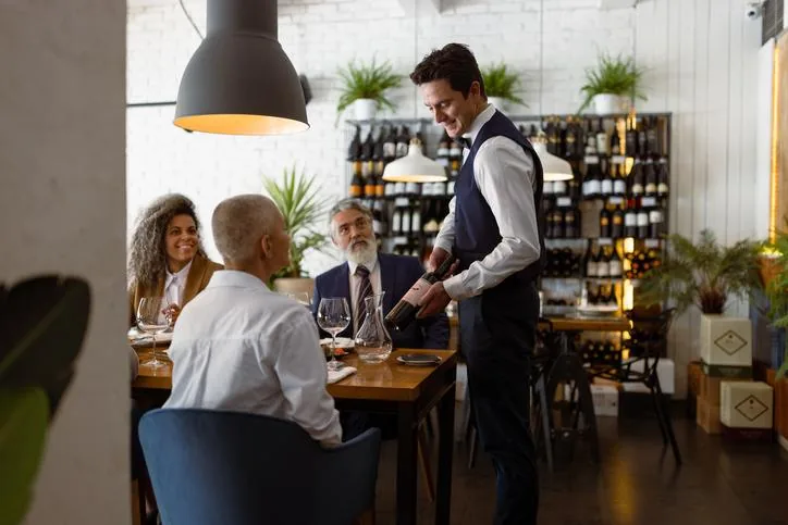 Camarero profesional presenta una botella de vino a comensales en una cena de negocios. Grupo de personas en un restaurante elegante con estanterías de vino al fondo.