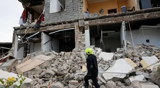 Un rescatista con casco amarillo camina entre los escombros de un edificio de varios pisos parcialmente colapsado.