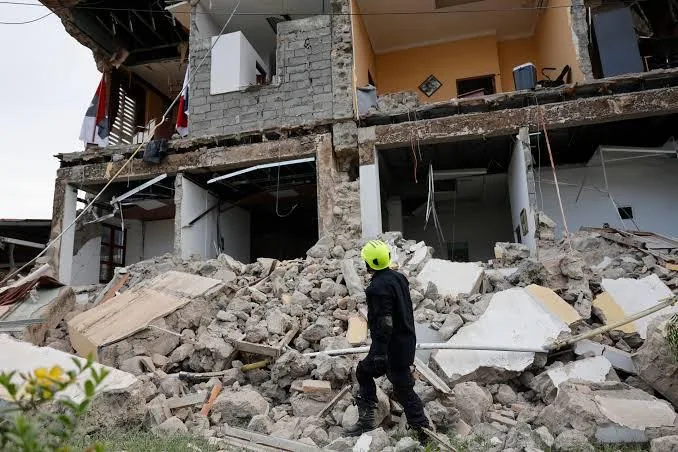 Un rescatista con casco amarillo camina entre los escombros de un edificio de varios pisos parcialmente colapsado.