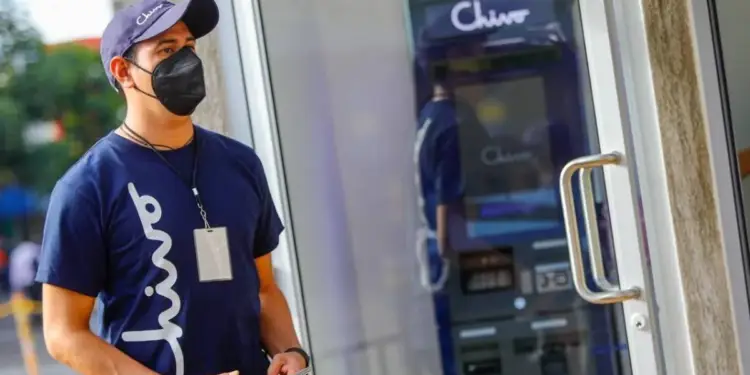 Hombre con mascarilla y uniforme azul con logo 'Chivo' frente a un cajero automático 'Chivo', sosteniendo un smartphone. Representa la interacción con tecnología financiera.