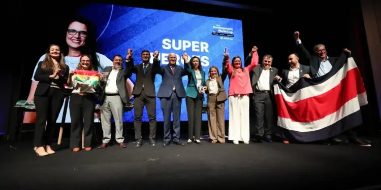 Un grupo de personas vestidas formalmente en un escenario, celebrando con banderas y trofeos. Al fondo, una pantalla muestra el texto 'SUPER HEROES'.