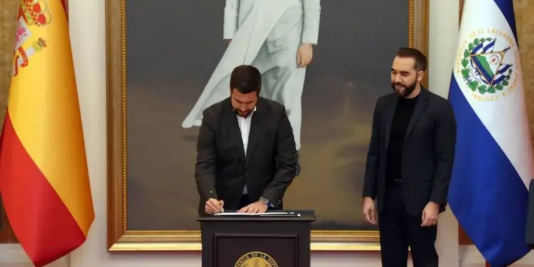 Dos hombres en trajes oscuros en un evento oficial. Uno firma documentos en un podio con el sello de El Salvador, el otro observa. Banderas de España y El Salvador flanquean la escena.