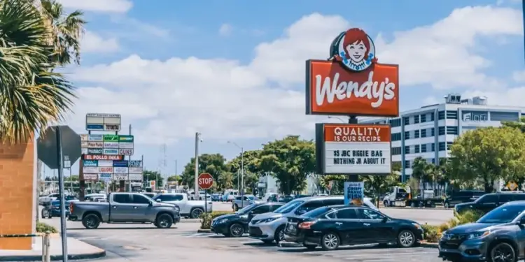 Vista exterior de un estacionamiento con un letrero de Wendy's, automóviles y varios negocios al fondo bajo un cielo azul.