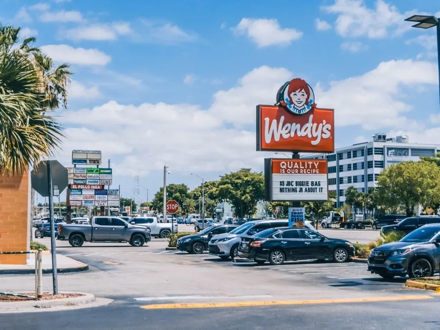 Vista exterior de un estacionamiento con un letrero de Wendy's, automóviles y varios negocios al fondo bajo un cielo azul.