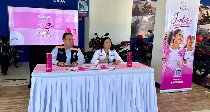 Dos personas sentadas en una mesa cubierta con mantel blanco, frente a una pantalla grande que muestra "UMA Yo Corro Por Ellas" y un banner rosa con mujeres corriendo y el logo "Movicancer Juntos corremos por ellas".