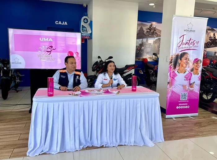 Dos personas sentadas en una mesa cubierta con mantel blanco, frente a una pantalla grande que muestra "UMA Yo Corro Por Ellas" y un banner rosa con mujeres corriendo y el logo "Movicancer Juntos corremos por ellas".