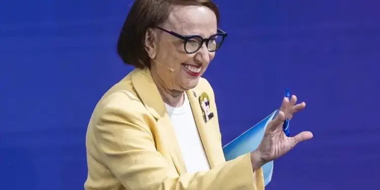 Mujer mayor sonriente con gafas y blazer amarillo, sosteniendo documentos y un bolígrafo, gesticulando en un evento profesional.