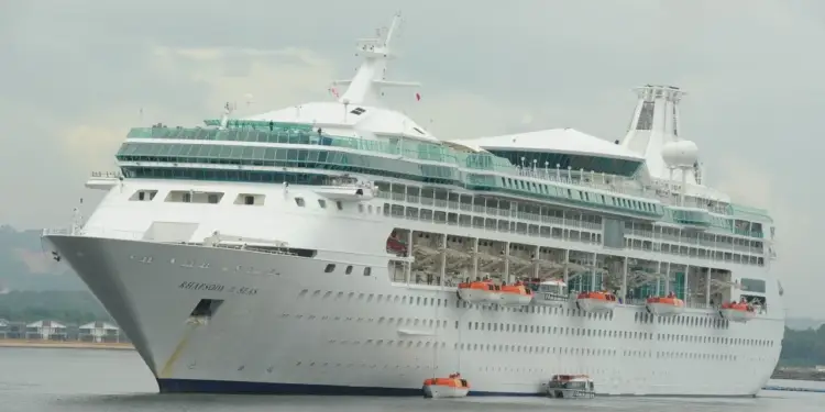 Gran crucero blanco 'Rhapsody of the Seas' en el agua, con múltiples cubiertas, ventanas y botes salvavidas naranjas. Se ven casas y árboles en la costa.