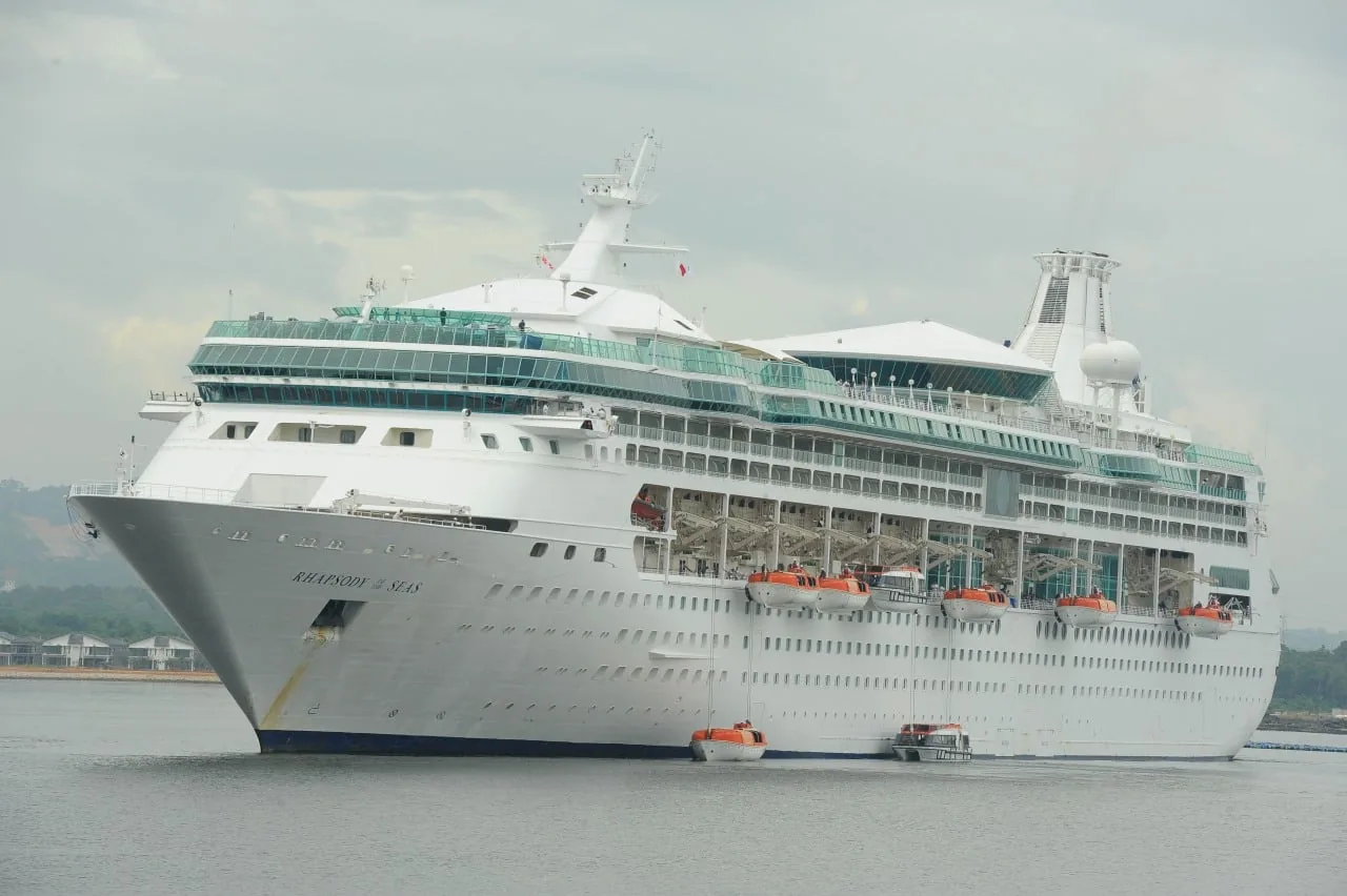 Gran crucero blanco 'Rhapsody of the Seas' en el agua, con múltiples cubiertas, ventanas y botes salvavidas naranjas. Se ven casas y árboles en la costa.