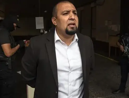 Hombre con traje oscuro y camisa blanca, con las manos atadas a la espalda, siendo observado por personal policial con equipo táctico.
