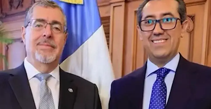 Dos hombres con traje formal y corbata posando juntos frente a una bandera y paneles de madera.