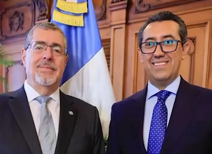 Dos hombres con traje formal y corbata posando juntos frente a una bandera y paneles de madera.