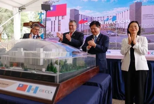 Grupo de funcionarios aplaudiendo junto a maqueta de hospital, con banderas de Taiwán y Guatemala en fondo, simbolizando cooperación.